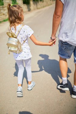 Bir baba güneşli bir günde kızının yanında yürür, sakin bir yolu keşfederken el ele tutuşur. Küçük kız küçük bir sırt çantası taşıyor ve gülümsüyor, neşeli bir anı paylaşıyor..