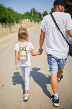 Bir baba güneşli bir günde kızının yanında yürür, sakin bir yolu keşfederken el ele tutuşur. Küçük kız küçük bir sırt çantası taşıyor ve gülümsüyor, neşeli bir anı paylaşıyor..