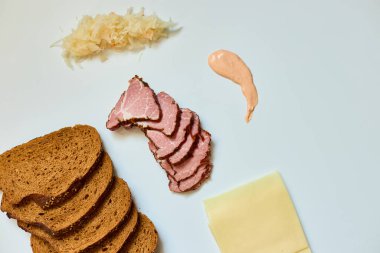 Dilimlenmiş çavdar ekmeği, konserve sığır eti, peynir, lahana turşusu ve sos beyaz bir yüzey üzerinde ustalıkla sergileniyor. 