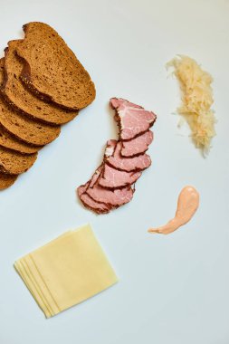 Dilimlenmiş çavdar ekmeği, konserve sığır eti, peynir, lahana turşusu ve sos beyaz bir yüzey üzerinde ustalıkla sergileniyor. 