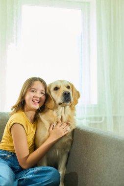 Neşeli bir genç kız rahat bir koltukta oturup Golden Retriever 'ına sarılıyor. Güneş ışığı büyük pencerelerden içeri akar ve oturma odasında sıcak, davetkar bir atmosfer yaratır..