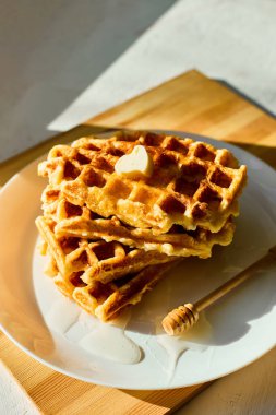 Beyaz bir tabağa istiflenmiş leziz waffle 'lar ve üzerinde küçük bir parça tereyağı eriyor. Yanlara serpiştirilmiş bal ekliyor tatlı bir dokunuş, hepsi de güneş ışığında güzelce aydınlatılıyor..