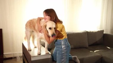 Kahverengi saçlı genç bir kız bir Golden Retriever 'a sarılıp kanepede dururken gülümsüyor. Işıl ışıl oturma odası, birlikte geçirdikleri mutlu ana sıcaklık katıyor..