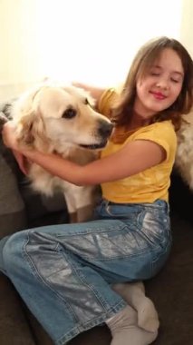 Kahverengi saçlı genç bir kız bir Golden Retriever 'a sarılıp kanepede dururken gülümsüyor. Işıl ışıl oturma odası, birlikte geçirdikleri mutlu ana sıcaklık katıyor..