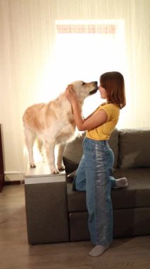 Kahverengi saçlı genç bir kız bir Golden Retriever 'a sarılıp kanepede dururken gülümsüyor. Işıl ışıl oturma odası, birlikte geçirdikleri mutlu ana sıcaklık katıyor..