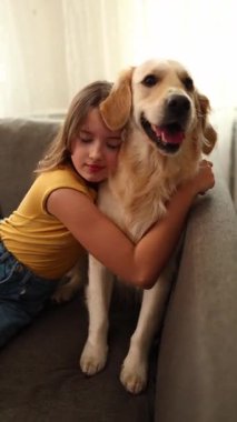 Kahverengi saçlı genç bir kız bir Golden Retriever 'a sarılıp koltukta otururken gülümsüyor. Işıl ışıl oturma odası, birlikte geçirdikleri mutlu ana sıcaklık katıyor..