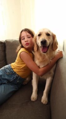Kahverengi saçlı genç bir kız bir Golden Retriever 'a sarılıp koltukta otururken gülümsüyor. Işıl ışıl oturma odası, birlikte geçirdikleri mutlu ana sıcaklık katıyor..