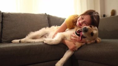 Neşeli bir genç kız, parlak bir oturma odasında yumuşak bir kanepede Golden Retriever 'ına sarılıyor. Güneş ışığı, birbirlerine eşlik ederek sıcak bir atmosfer yaratıyor..