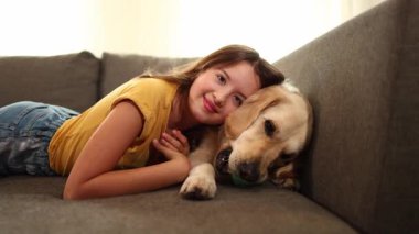 Genç bir kız kanepede uzanmış Golden Retriever 'ıyla şakalaşıyor. Güneş ışığı odayı aydınlatır, sıcak ve davetkar bir atmosfer yaratır..