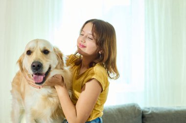 Genç bir kız, Golden Retriever 'ıyla doğal ışıkla dolu parlak, rahat bir oturma odasında kaliteli vakit geçirmekten zevk alır. Birlikte neşeli bir anı paylaşıyorlar..