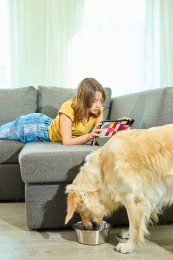 Genç bir kız tabletine odaklanmış bir şekilde kanepede yüzüstü yatıyor. Golden Retriever yerde bir kaseden yemek yiyor. Oturma odasında sıcak, sade bir atmosfer yaratıyor..