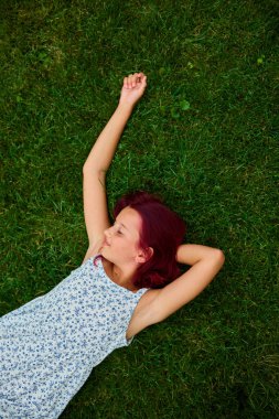 Canlı kızıl saçlı bir kız yemyeşil çimenlerin üzerinde uzanıyor, açık hava alanının sakinliğinin tadını çıkarıyor. Güneş ışığı, rahatladığında çiçekli elbisesini aydınlatıyor..