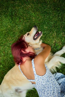 Kızıl saçlı bir kız Golden Retriever 'ıyla çimenlerde uzanıyor ve bir anlık sevgi paylaşıyor. Açık hava manzarası canlı ve sıcak, sevgi dolu bir bağ sergiliyor..