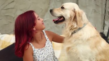 Çiçekli bir elbise giyen bir kız rahat bir salıncakta oturur ve Golden Retriever 'ına mutlu bir şekilde sarılır. Güneş batıyor, sıcak ve neşeli bir atmosfer yaratıyor..