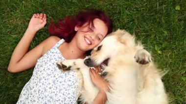 Genç bir kız çimenlerde uzanır, Golden Retriever köpeğiyle oynarken ışıl ışıl gülümser. Güneş parlıyor, neşe ve arkadaşlıkla dolu sıcak bir atmosfer yaratıyor..