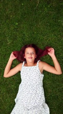 Uçan kızıl saçlı genç bir kız çiçek desenli bir elbise giyerek yemyeşil çimenlerin üzerine uzanır. Mutlu bir şekilde gülümser ve güzel bir günde sıcak güneş ışığının tadını çıkarır..