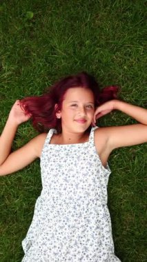 Uçan kızıl saçlı genç bir kız çiçek desenli bir elbise giyerek yemyeşil çimenlerin üzerine uzanır. Mutlu bir şekilde gülümser ve güzel bir günde sıcak güneş ışığının tadını çıkarır..