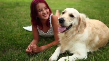 Kızıl saçlı bir kız yanında Golden Retriever 'la çimlerin üzerinde rahatlıyor. Yerel bir parkta güneşli bir günün tadını çıkarıyorlar. Arkadaşlıklarla dolu neşeli bir atmosfer yaratıyorlar..