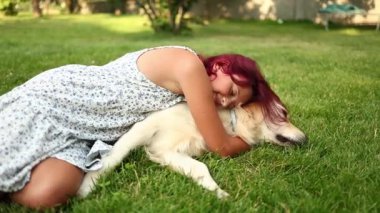 Genç bir kız dost canlısı Golden Retriever 'ına sarılırken yeşil çimlerde dinleniyor. Güneşli bir öğleden sonra bahçede neşeli ve sevgi dolu bir bağ paylaşıyorlar..