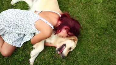 Genç bir kız dost canlısı Golden Retriever 'ına sarılırken yeşil çimlerde dinleniyor. Güneşli bir öğleden sonra bahçede neşeli ve sevgi dolu bir bağ paylaşıyorlar..