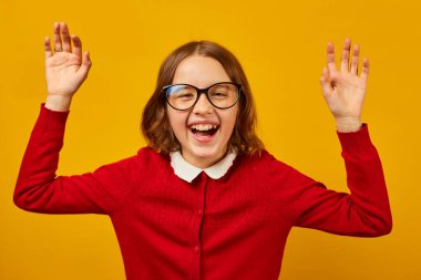 Kıvırcık saçlı neşeli bir kız heyecanla gülümserken ellerini kaldırır. Yuvarlak gözlükler takıyor ve kırmızı bir kazak giyiyor, mutluluk dolu canlı bir sahne yaratıyor..