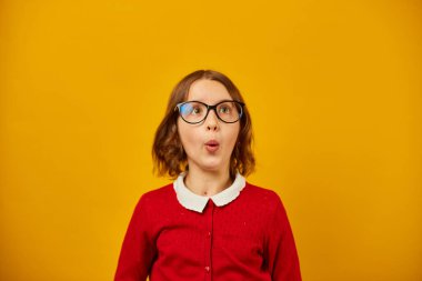 Gözlük takan genç bir kız ve kırmızı bir hırka parlak sarı bir duvara karşı neşeyle poz veriyor. İzleyicilerle iletişim kurarken ifadesi heyecan ve güven gösteriyor..