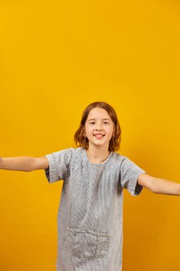 Genç bir kız kolları açık gülümsüyor ve başparmaklarını kaldırıyor. Çizgili bir gömlek giyer ve canlı sarı bir arka plana yaslanır. Mutluluk ve coşku saçar..