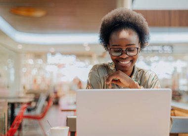 Genç bir Afro-Amerikalı kadın, alışveriş merkezinde kahve içerken laptopuyla video konferansı yapıyor..