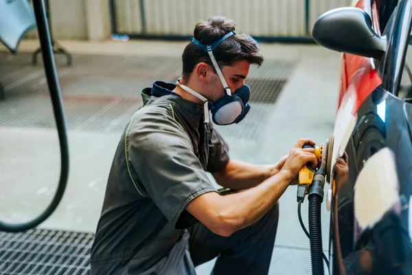 A young mechanic wearing a mask sands the door of a car before painting it.