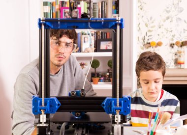 Bir öğrenci ve 3D yazıcıyla kameraya bakan genç bir öğretmen..