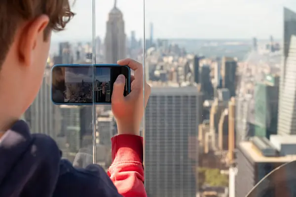 An unrecognizable teenager takes a cell phone photo of the New York City skyline on a sunny summer morning.