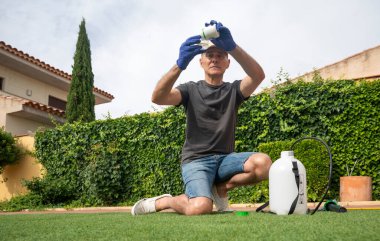 Koruyucu eldivenleri olan orta yaşlı bir adam bahçede böcek ilacı spreyi hazırlıyor.