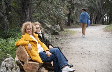 Yaşlı kadınlar bir orman parkında ahşap bir bankta otururken genç bir adam yürüyüp gidiyor.