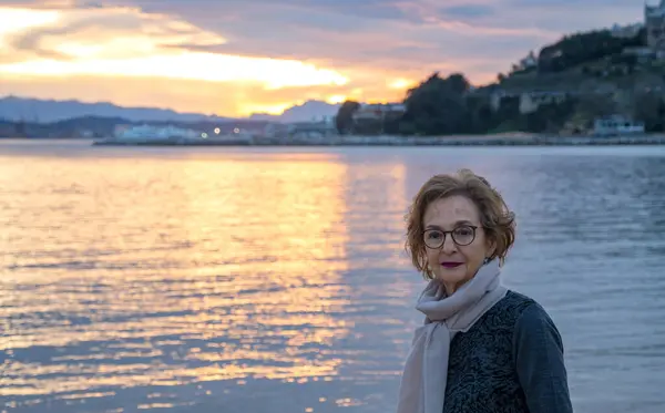 Güneş batarken suyun kenarında duran gözlük ve eşarp takan yaşlı bir kadın.