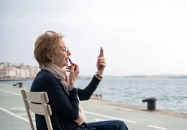Deniz kenarında cep telefonunu ayna olarak kullanıp ruj süren güzel bir son sınıf öğrencisi.