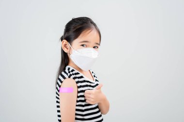 Tıbbi maskeli küçük bir Asyalı kız kolunu virüs aşısı sonrası bandajla gösteriyor. Çocuk ve çocuk aşılama kampanyası. Çocuğunu aşı konseptiyle koru.