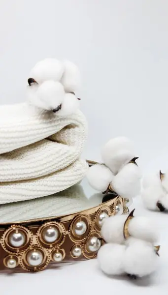 Stack of white knitted blankets folded and decorated with cotton sprigs with open bolls on gold tray with pearls.