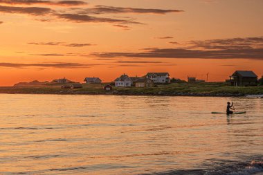 Lofoten, Norveç - 31 Temmuz 2023: Ramberg, Lofoten, Norveç yakınlarında kano süren bir kadın, sıcak bir yaz gecesi, arka planda evler, turuncu gün batımı, parçalı bulutlu.
