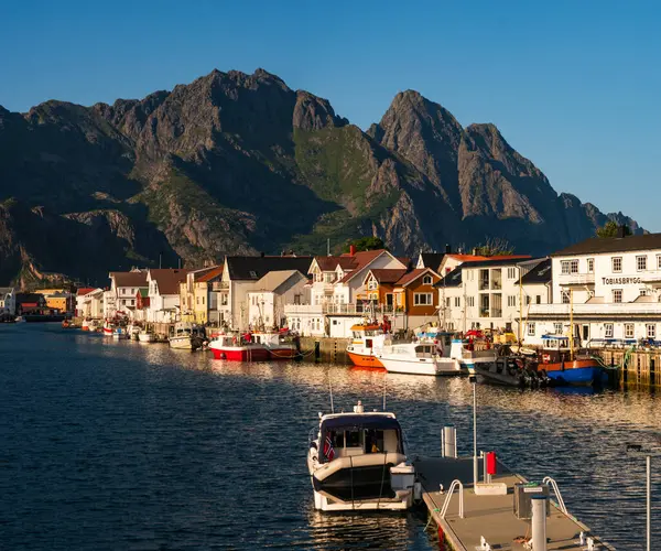 Lofoten, Norveç - 28 Temmuz 2023: Lofoten 'in güney kıyısındaki küçük Henningsvaer köyü, güneş ışığı, beyaz evler ve dağ tepeleriyle dolu bir yaz akşamı,.