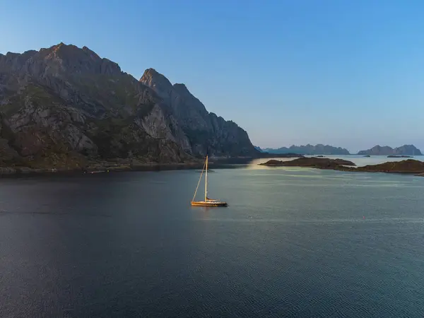 stock image A sailboat under the midnight sun, behind a large hill in Lofoten, near Henningsvaer, in the distance tall mountains, calm sea.