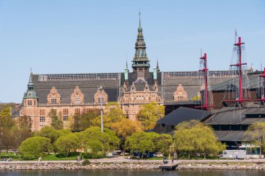 İsveç 'in Stockholm kentindeki Skeppsholmen Adası' ndan güneşli bir bahar gününde görülen İskandinav Müzesi ve Vasa Müzesi. Mavi gökyüzü ve yeşil ağaçlar. İnsanlar suyun kenarında dinleniyor.