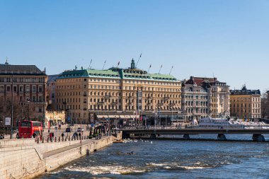 Stockholm - 30 Mart 2024 Stockholm ile şehir merkezi arasındaki nehir. Grand Otel, İsveç bayrakları, Strmbron Köprüsü. Parlak güneş ışığı, bahar. Mavi gökyüzü.