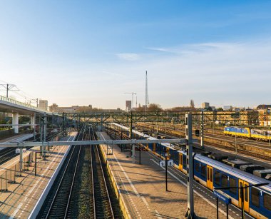 Den Haag, Hollanda - 25 Mart 2024: Lahey 'in merkez tren istasyonunda sabah güneşinde raylar ve trenler. Mavi gökyüzü. Yoldan geçen banliyö trenleri.