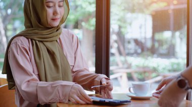 Genç, güzel, Asyalı Müslüman bir kadın güneşli bir günde kahve dükkanında çalışırken ve cep telefonuyla oynarken dinleniyor.