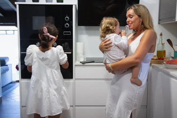 Young mother in the kitchen with baby in her arms happy with her other older daughter