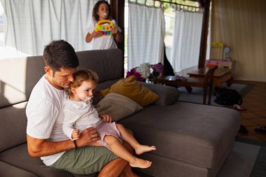 Baba ve kızları oturma odasında, biri oynuyor ve ufaklık giyiniyor..