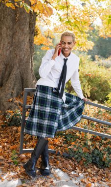 Parktaki genç adam cep telefonuyla konuşurken gülümsüyor İskoç eteği giymiş, dikey bir fotoğrafla.