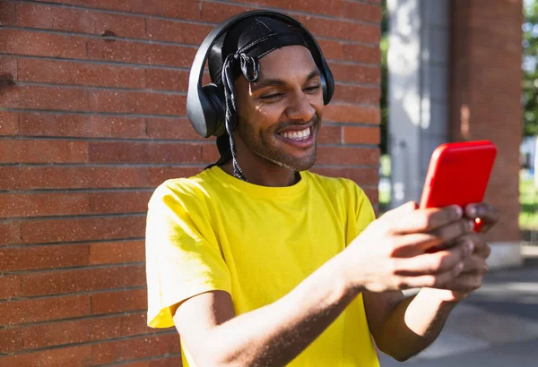 Young black man smiling happily with headphones smiling acting with mobile phone in the street, copy space