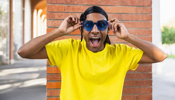 Beautiful young black man funny with cheerful expression wearing blue glasses, looking at camera in the street