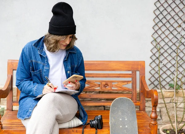 A thoughtful urban artist sits on a wooden bench, immersed in her sketchbook and smartphone, with a skateboard and camera nearby, epitomizing a creative lifestyle.
